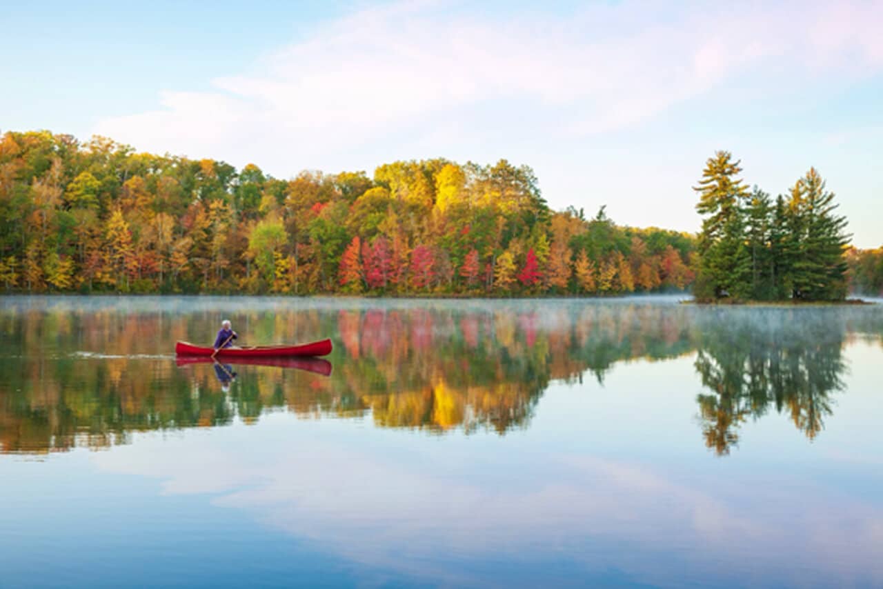 A man paddles a canoe on a serene lake, framed by vibrant fall foliage reflecting in the water.