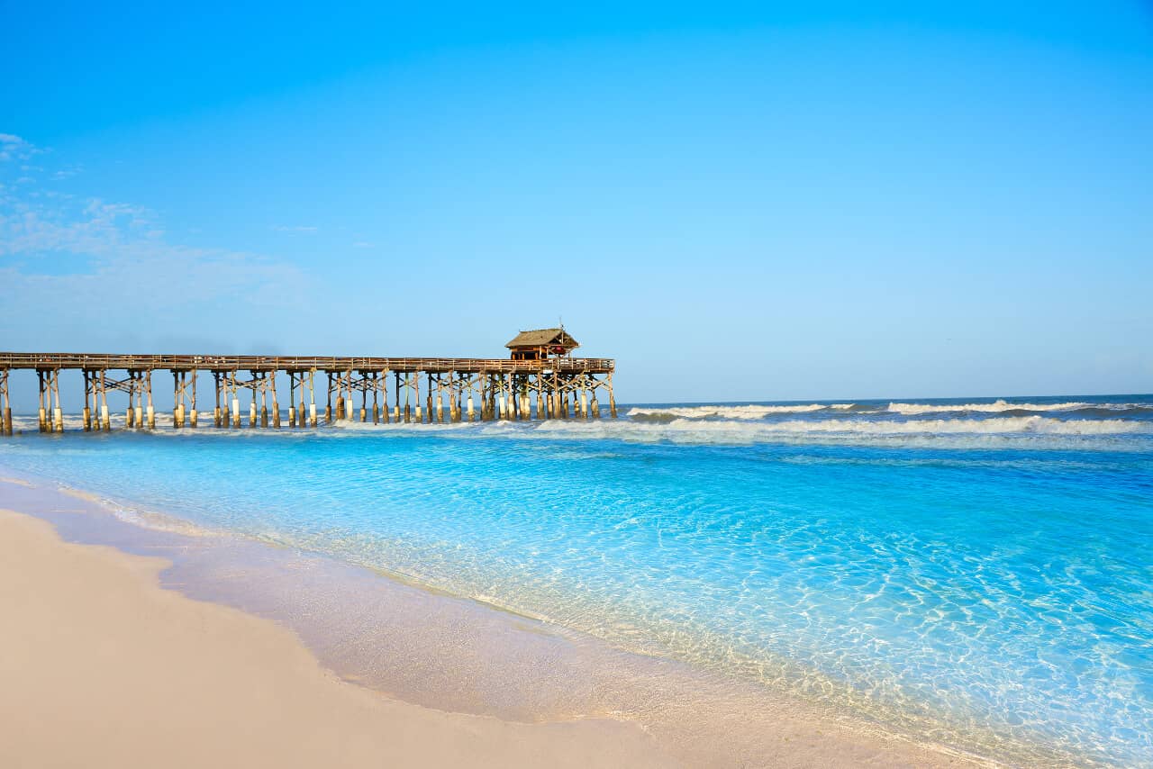 Wooden pier extends over turquoise ocean water with gentle waves, under a clear blue sky. Sandy beach in the foreground.